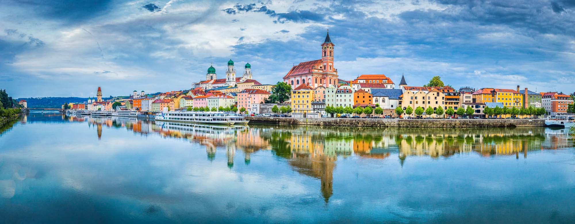 Passau Reflected in the Danube River A panoramic view of the historic Bavarian city of Passau with its iconic colorful buildings and church spires reflected perfectly in the calm Danube River during golden hour. The picturesque riverside town showcases traditional Central European architecture with vibrant pastel-colored facades and a prominent red-roofed cathedral, creating a stunning mirror image on the water.
