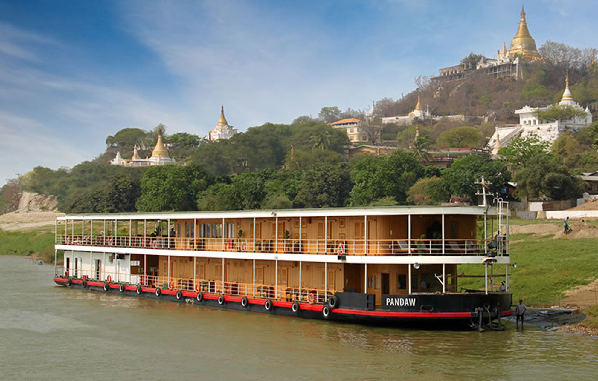 A two-deck river cruise vessel named Pandaw moored on the Irrawaddy River in Myanmar, with golden pagodas and Buddhist temples visible on the hillside in the background under clear blue skies.