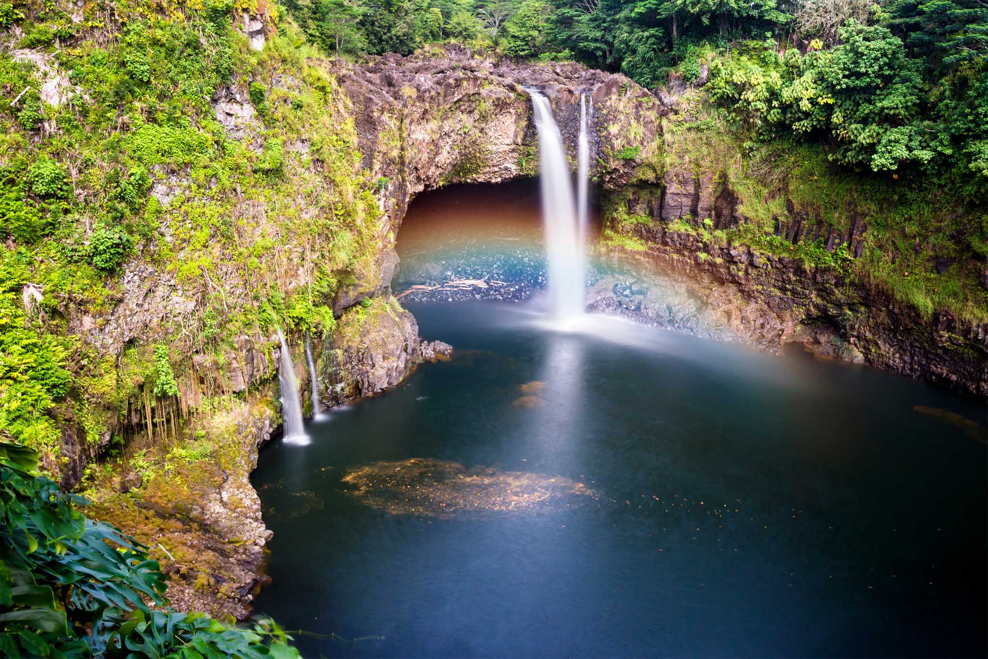 A stunning waterfall cascades into a deep natural pool surrounded by lush green vegetation and moss-covered basalt cliffs in Hilo, Hawaii. The pristine turquoise waters and verdant landscape create a serene tropical paradise, with the waterfall's misty spray catching sunlight to create subtle rainbow effects.