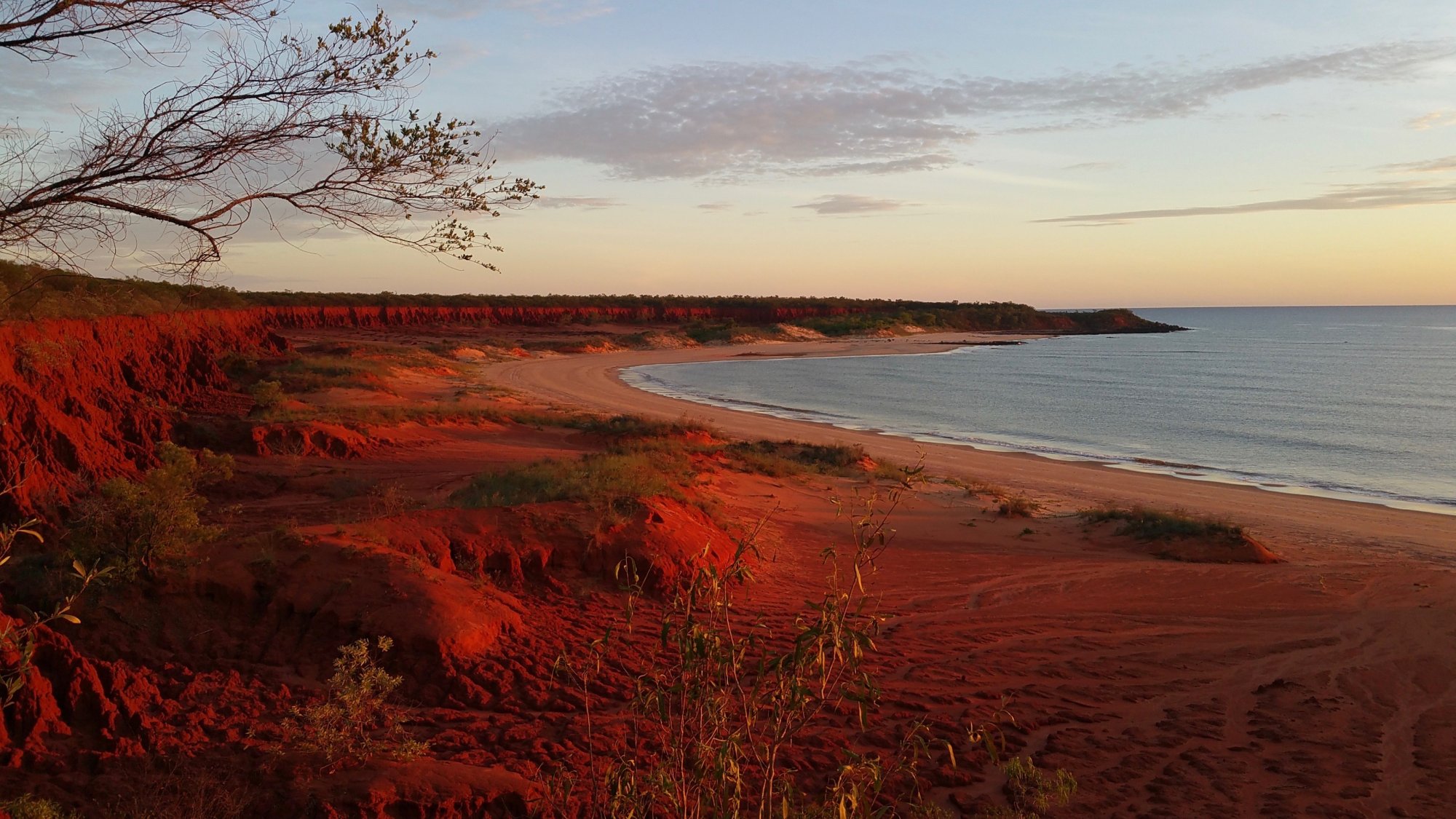 A stunning coastal landscape featuring dramatic red sandstone cliffs overlooking a curved sandy beach and calm waters at dusk. The warm golden light of sunset illuminates the distinctive rust-colored earth formations, creating a striking contrast with the blue-grey ocean and pale sky beyond.