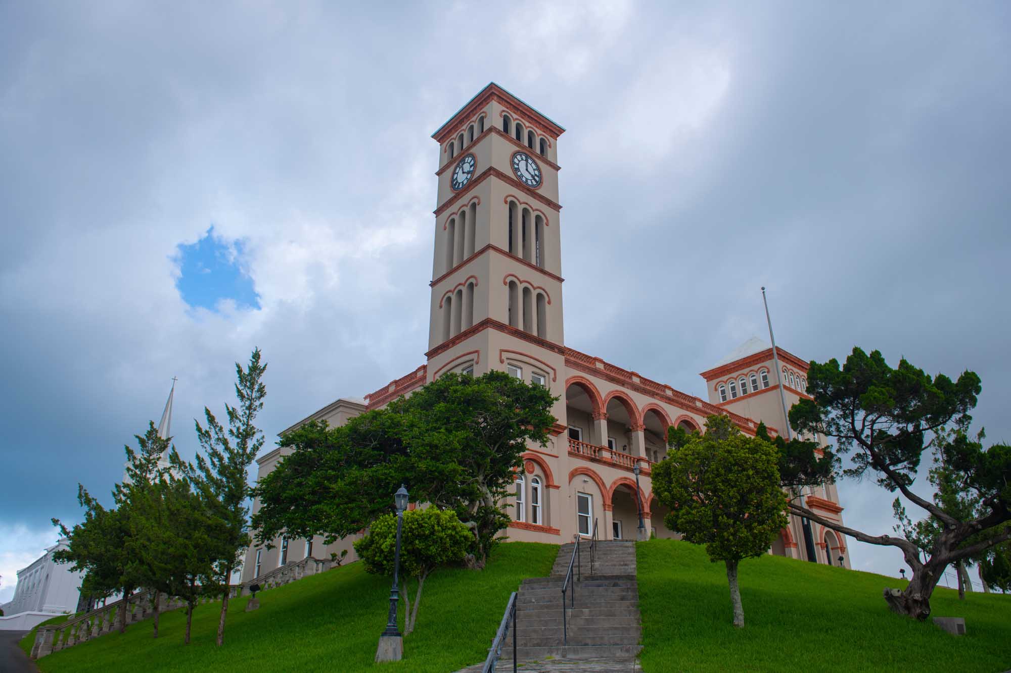 A distinctive pink and cream-colored colonial government building featuring a prominent clock tower, situated on a manicured green hillside in Hamilton's city center.