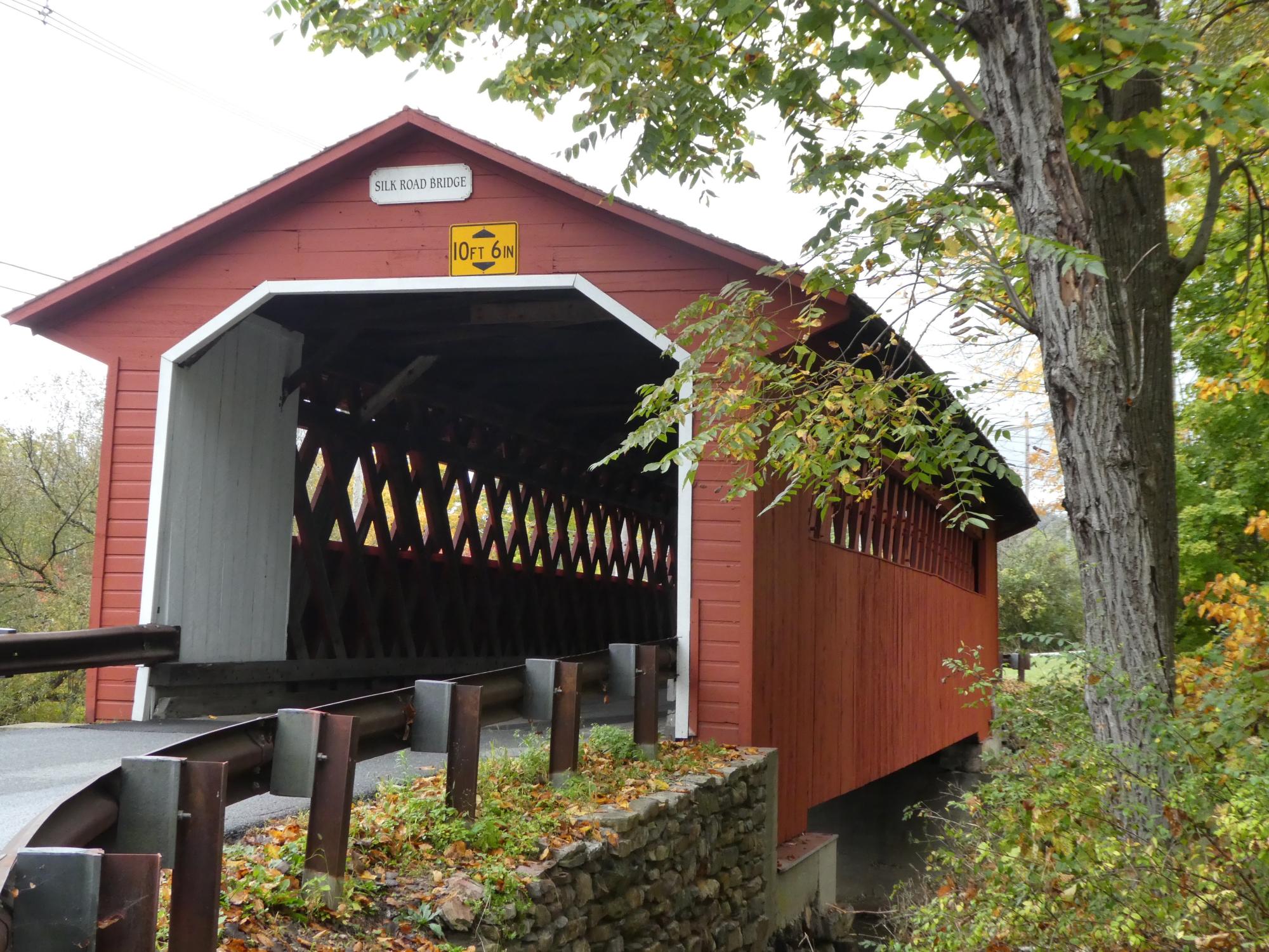 The historic Silk Road covered bridge, a red wooden structure with white trim and distinctive latticed interior, spanning across a stone foundation. The bridge features a clearance height of 10 feet 6 inches and is surrounded by autumn foliage and mature trees.