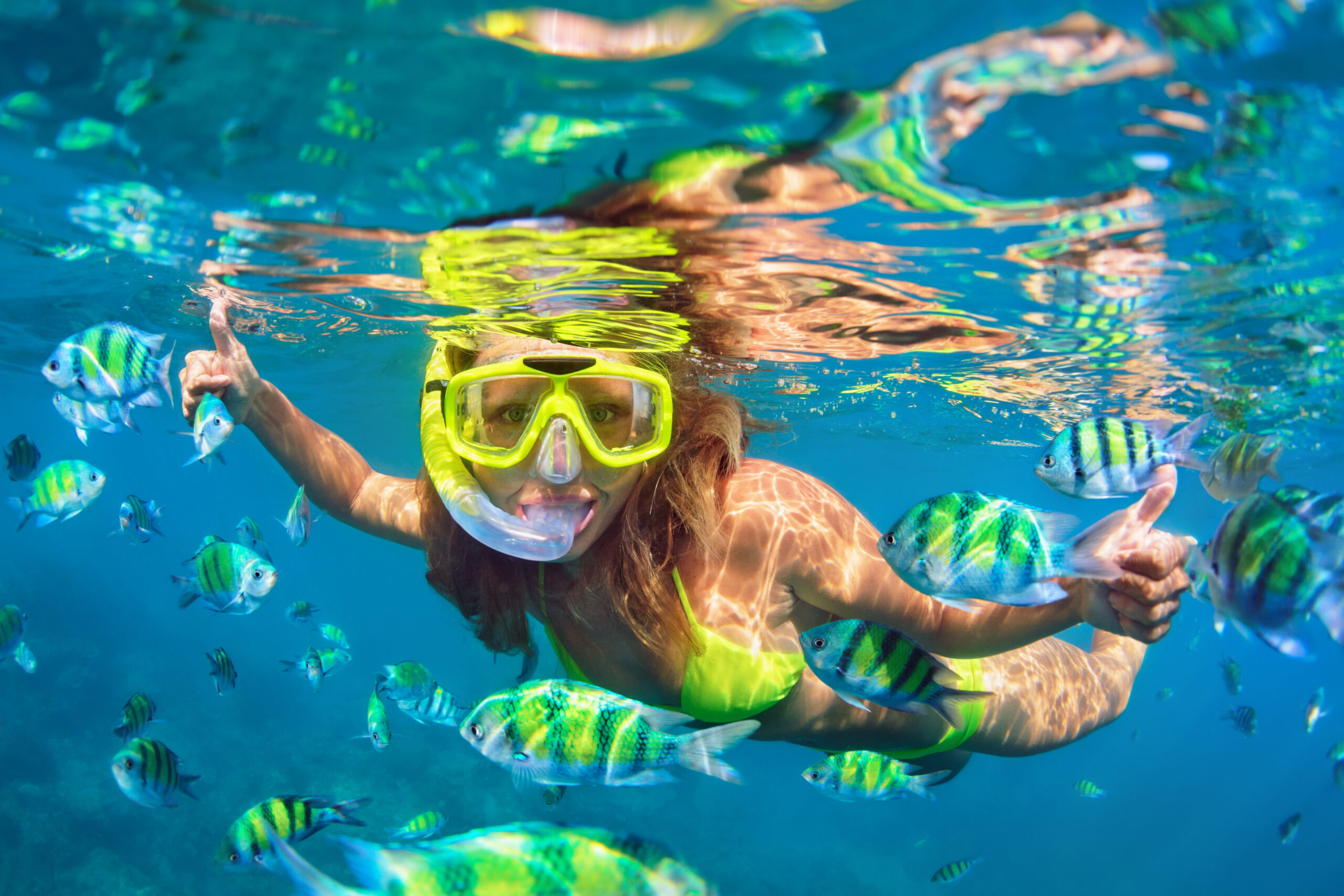 Snorkeling with Tropical Fish in Clear Blue Waters A young woman snorkels in crystal-clear tropical waters wearing a bright yellow-green snorkel mask and matching swimwear, surrounded by vibrant striped tropical fish with yellow and blue markings. She smiles at the camera with her arms outstretched, capturing the joy and wonder of underwater exploration in a pristine marine environment.