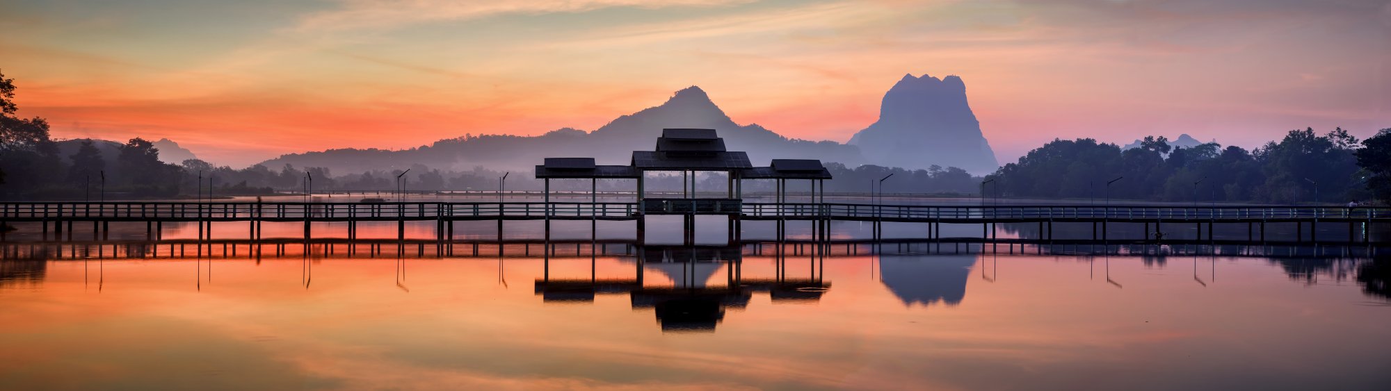 A serene sunrise scene over a calm lake featuring a traditional wooden pavilion structure with distinctive tiered roofing, silhouetted against dramatic mountain peaks in the background. The tranquil water perfectly mirrors the vibrant orange and pink sky, creating a symmetrical composition that evokes peaceful Southeast Asian landscapes.