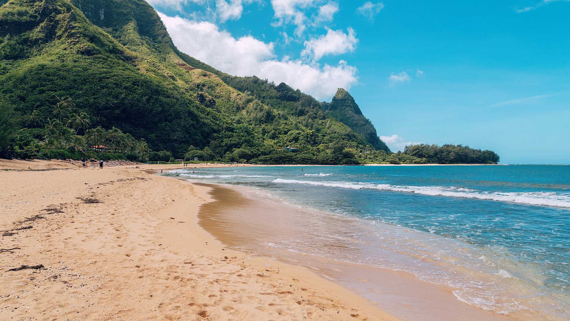 A pristine sandy beach with turquoise waters and gentle waves, framed by dramatic lush green cliffs and mountains. The idyllic tropical setting features a small number of visitors enjoying the shoreline beneath a bright blue sky with scattered clouds.