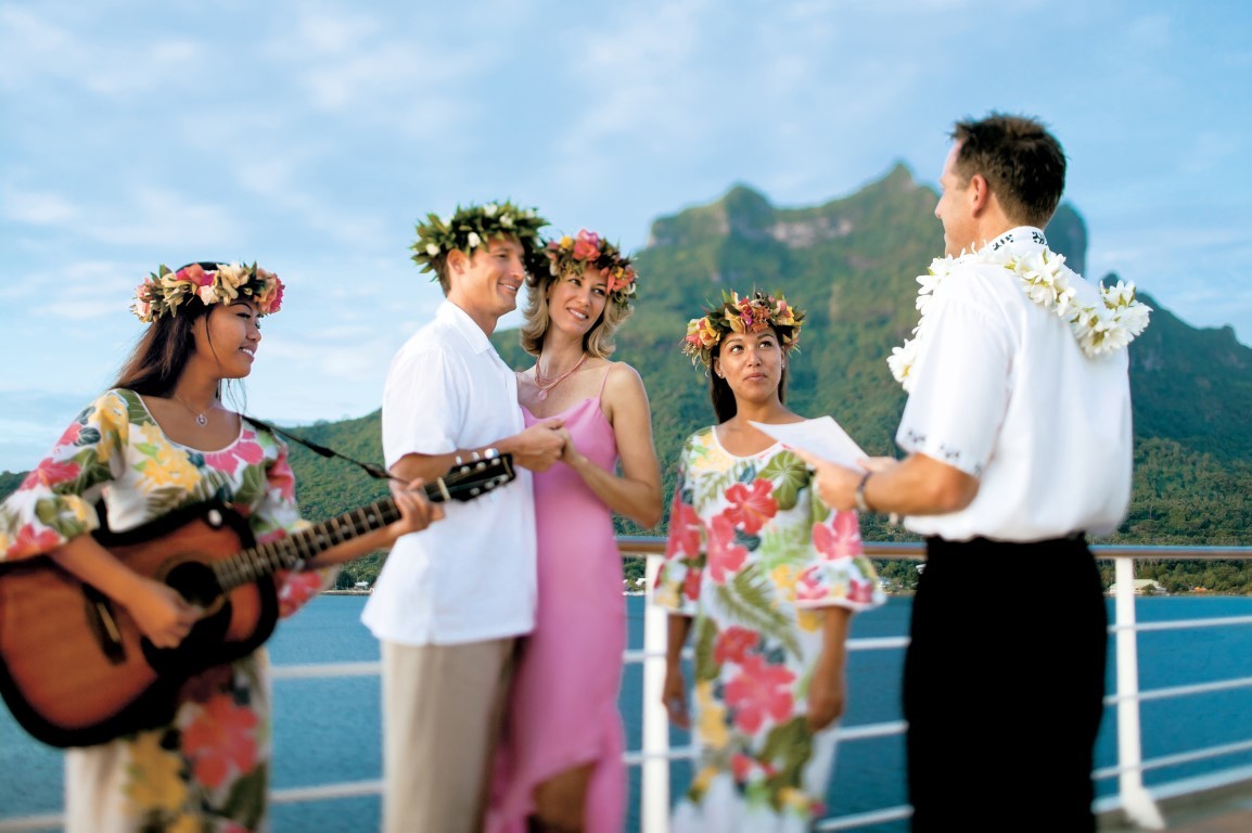 Tropical Island Wedding Celebration on Ship A joyful wedding celebration on a cruise ship deck featuring five people in tropical attire and flower garlands, with a lush green volcanic island and turquoise ocean visible in the background.