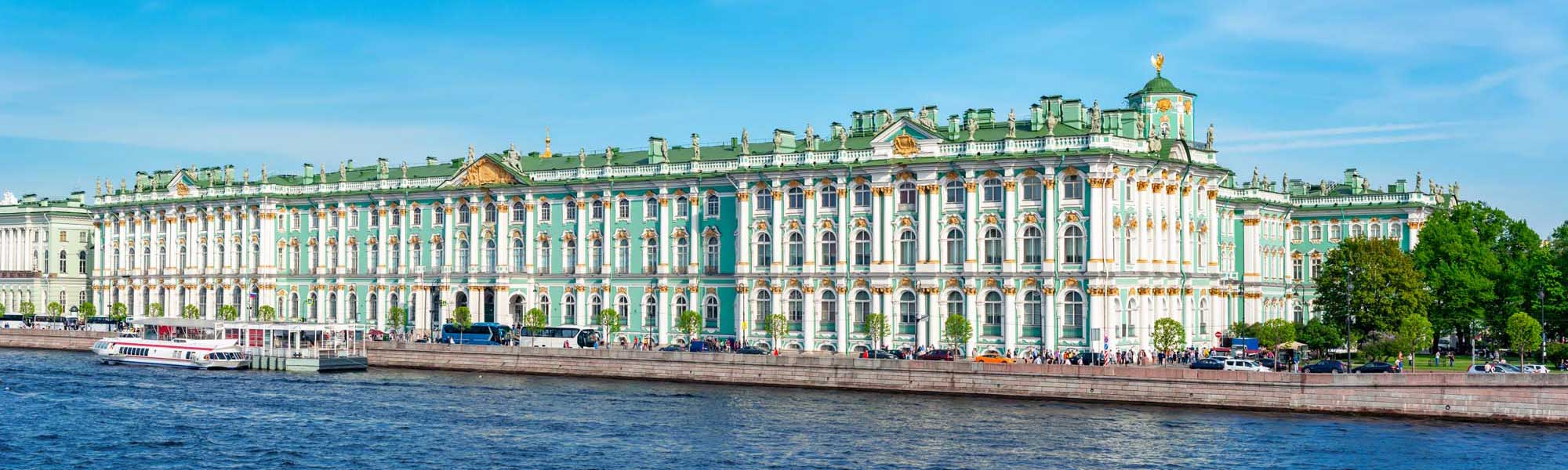 Winter Palace on the Neva River The magnificent Winter Palace in St. Petersburg, Russia, displaying its iconic turquoise and white Baroque facade with golden ornamental details, as viewed from the Neva River. The grand 18th-century imperial residence features multiple stories of arched windows, decorative moldings, and a distinctive green roof punctuated by ornamental spires.