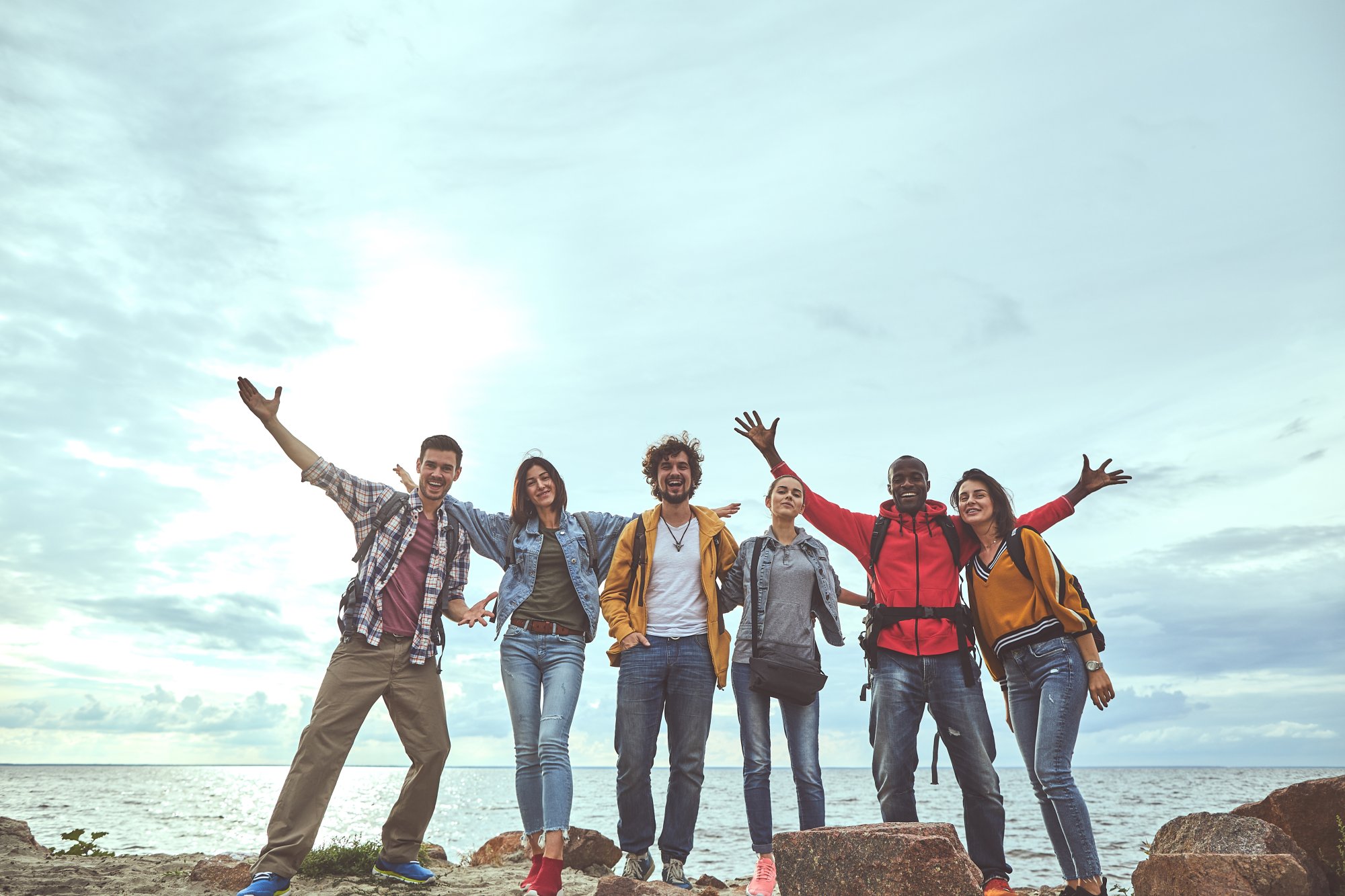 adobestock-223985854 A diverse group of six young adults standing on rocks by the ocean with their arms raised in celebration and joy, capturing a moment of happiness and camaraderie during a travel outing.