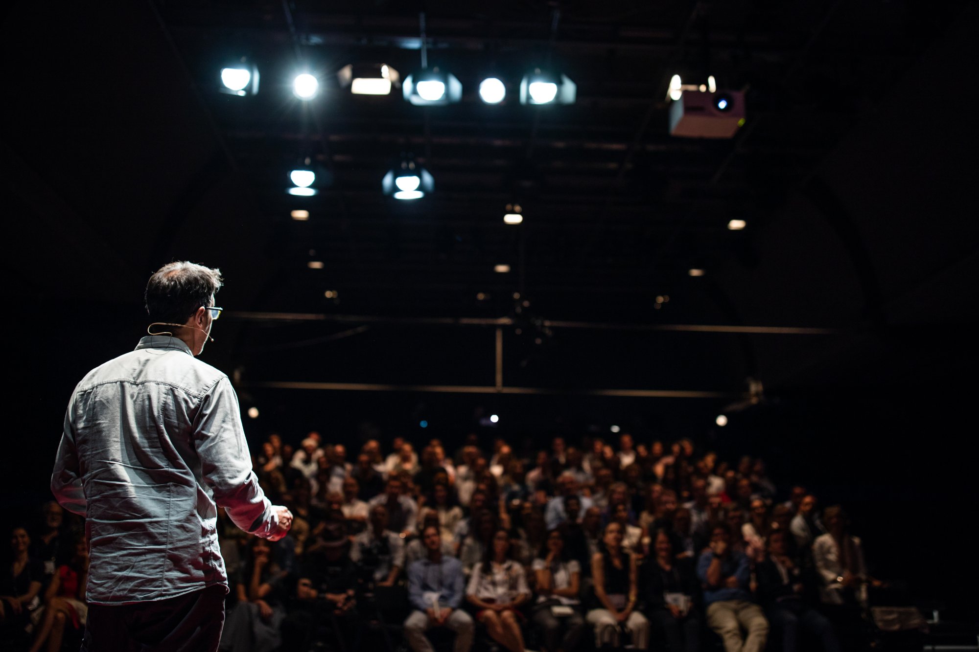 adobestock-224515728 A speaker addressing a large audience in a darkened theater, with professional stage lighting and an engaged crowd seated in the foreground.