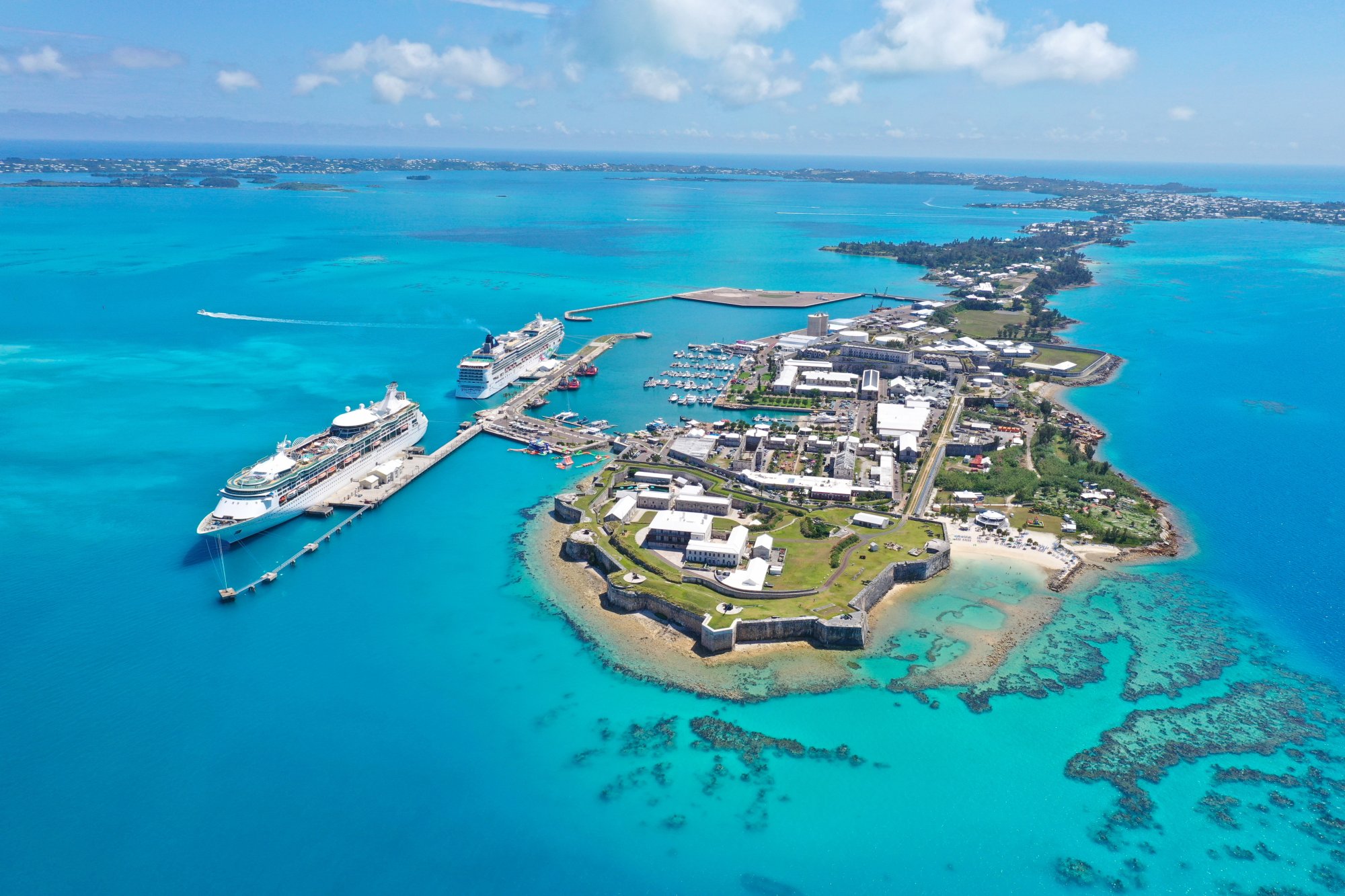 An aerial view of a large cruise ship docked at King's Wharf in Bermuda, showcasing the iconic fortress and colonial architecture alongside turquoise waters.