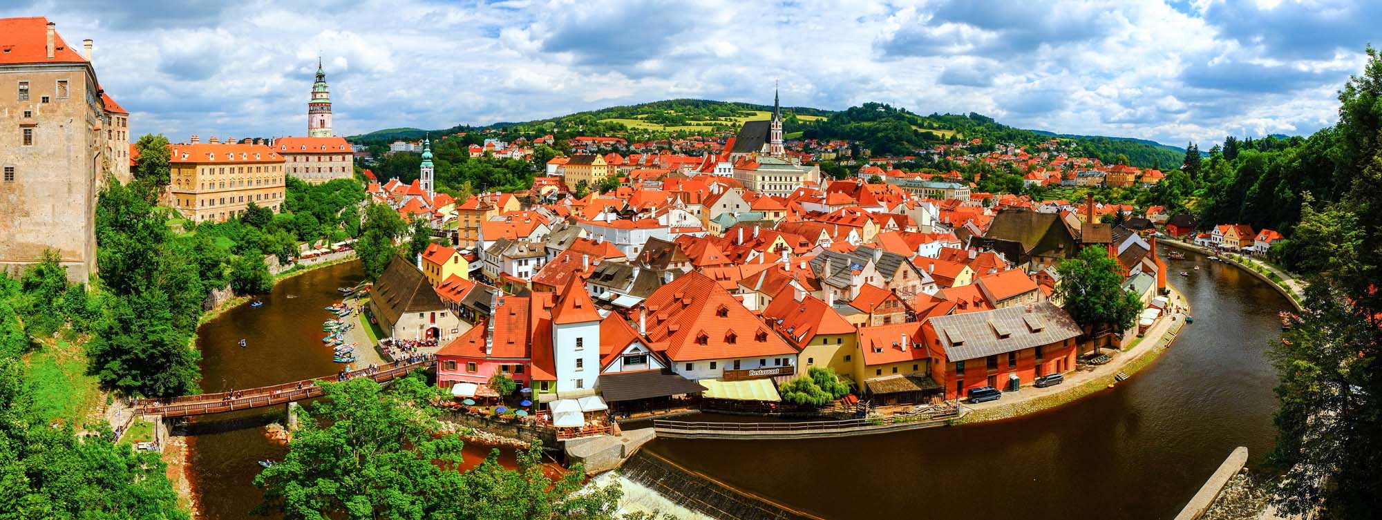 Český Krumlov Historic Town Panorama A stunning panoramic view of Český Krumlov, a UNESCO World Heritage Site in the Czech Republic, featuring the iconic red-tiled roofs of the medieval old town nestled within a dramatic bend of the Vltava River, with the historic castle and church spires rising prominently against forested hills under a partly cloudy sky.