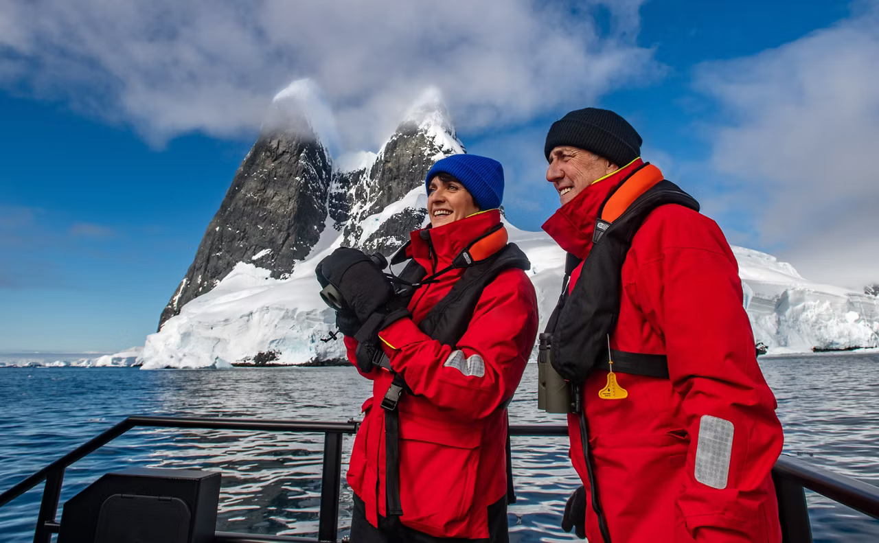 Two expedition-dressed travelers in bright red parkas stand on the deck of a zodiac or small