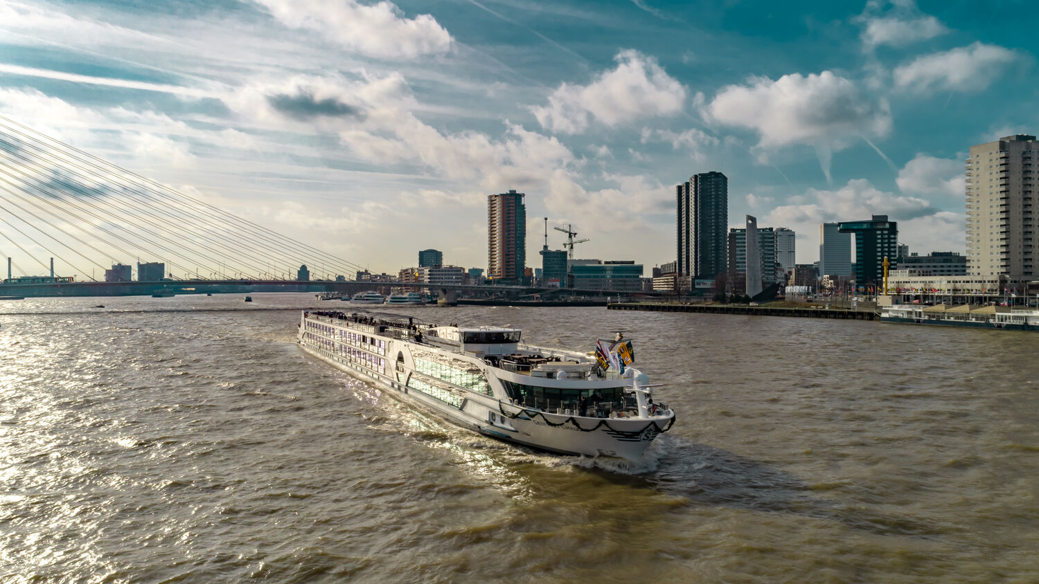 A river cruise ship, the Geoffrey Chaucer, navigates a wide European waterway with modern high-rise