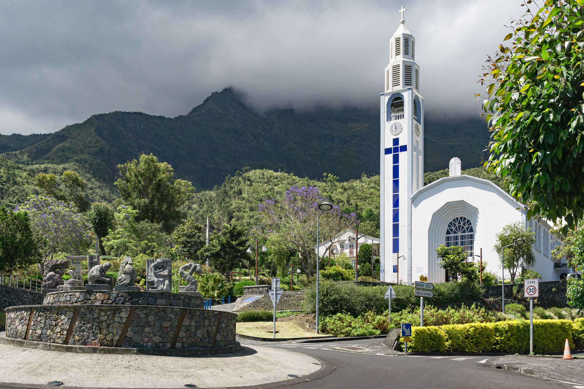 A modern white church with a tall bell tower featuring a prominent blue cross stands in a lush