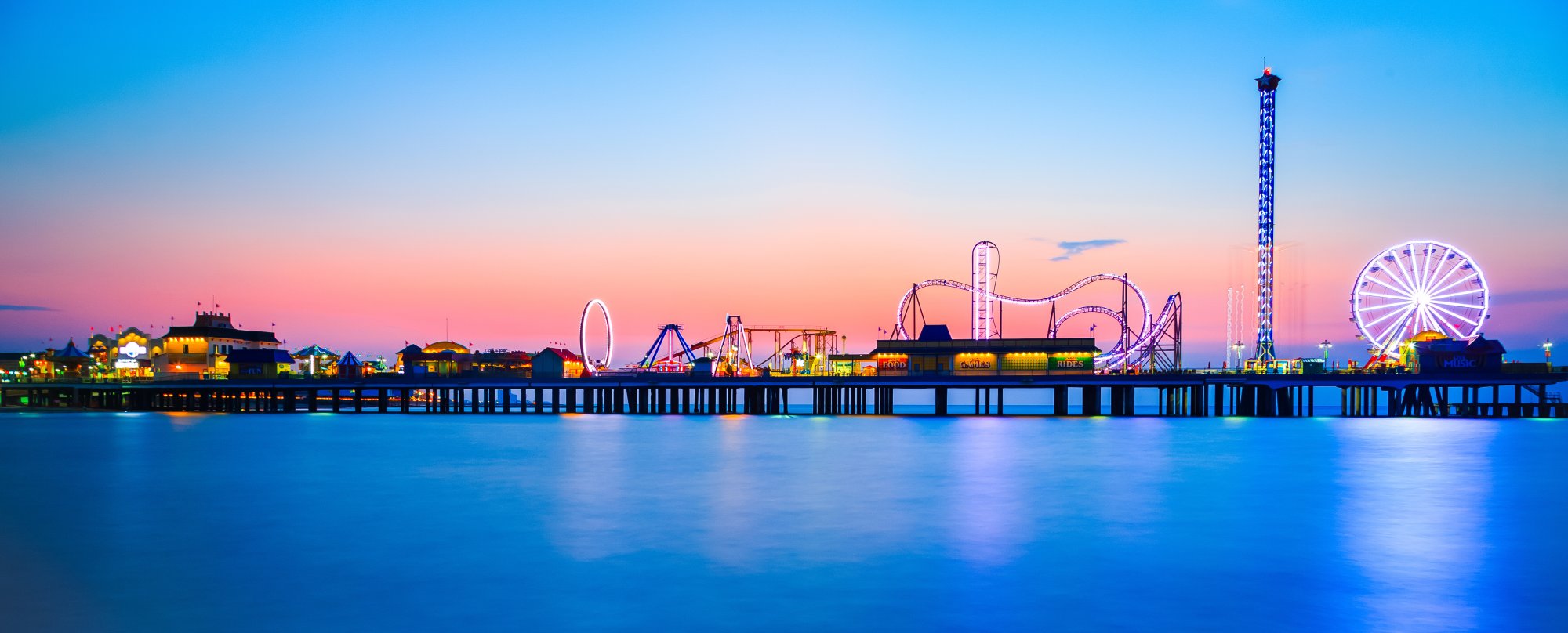 Galveston Pleasure Pier at Twilight An illuminated amusement pier extending into calm blue waters at dusk, featuring a Ferris wheel